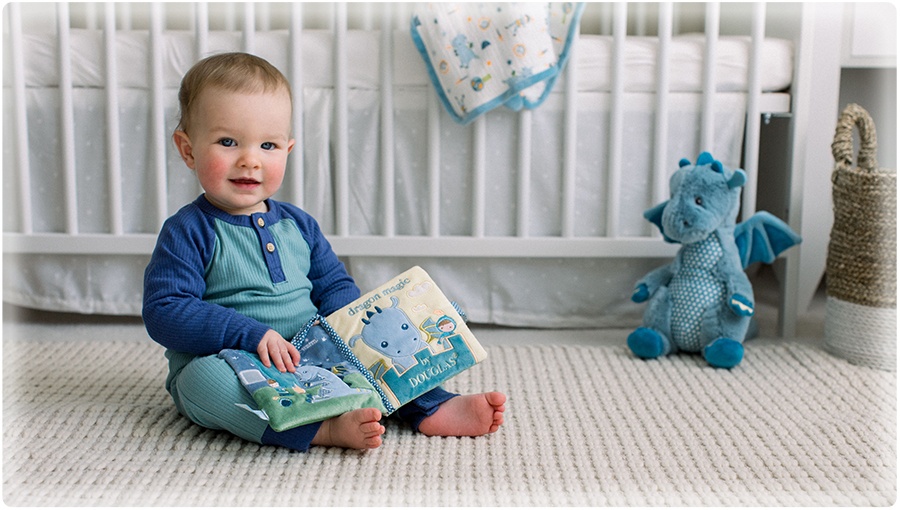 A smiling baby boy dressed in a blue romper sits on the floor in front of a white crib. He holds the soft, plush activity book from the DOUGLAS Baby Demitri Dragon character collection. Behind him a coordinating Blanki Lovey blanket is draped over the edge of the crib and the Demitri Dragon Plumpie stuffed animal sits near him on the floor.