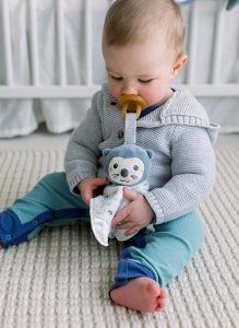 A baby boy sits on the floor holding the DOUGLAS Baby Indy Otter Paci Lovey. Made of cotton muslin and plush, the Paci Lovey is affixed to the little boy's pacifier and provides soft, silky textures that will sooth and comfort.
