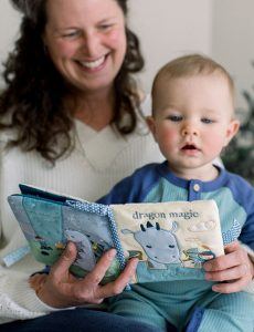 A mother sits with her infant son on her lap while reading a soft, plush activity book titled "Dragon Magic" that features DOUGLAS' Demitri Dragon character.