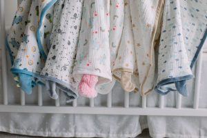 A selection of five different DOUGLAS Baby Blanki Lovey blankets hang draped over the edge of a white crib. Their cotton muslin fabric is trimmed with plush for a decadently soft feel. 