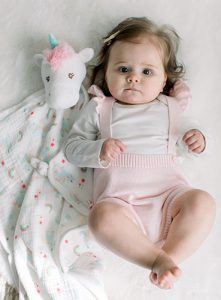 A baby girl wearing a pastel pink and white outfit lies on her back next to the Blanki Lovey blanket from the DOUGLAS Baby Emilie Unicorn character collection. The Blanki Lovey is made with cotton muslin and features a soft, plush face.