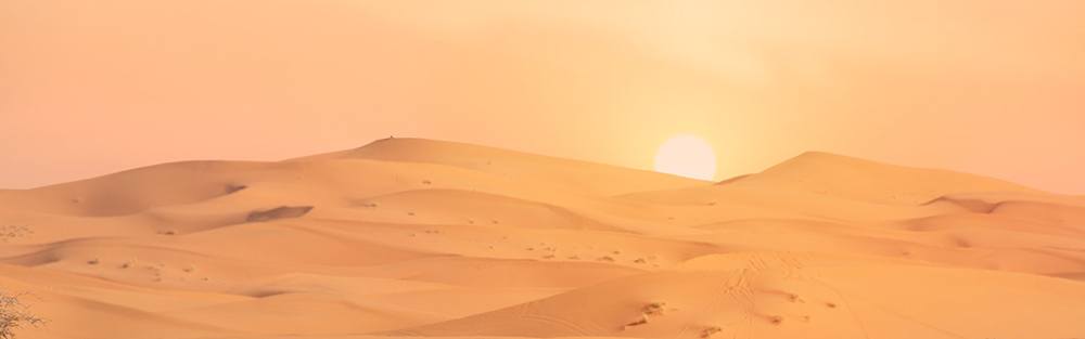 The glowing orb of the sun shines over an assortment of sandy desert dunes.